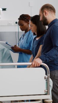 Physician pediatric doctor monitoring disease symptoms while african american nurse checking IV drip bag in hospital ward. Sick little kid resting in bed recovering after medicine surgery