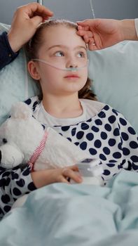 Worried father sitting with hospitalized sick daughter during medical examination in hospital ward. Specialist pediatrician doctor monitoring temperature to hospitalized child with breathing disease