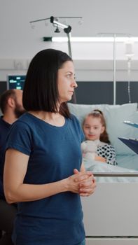 Mother discussing medical treatment with pediatrician doctor during disease examination in hospital ward. Girk kid resting in bed while african american nurse writing sickness diagnosis on clipboard