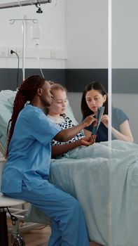 African american nurse showing lungs radiography explaining breathing disease symptoms discussing recovery treatment. Sick little chid resting in bed during clinical examination in hospital ward