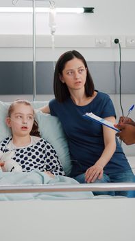 African american pediatric nurse explaining recovery treatment to worried parents monitoring sickness symptoms. Sick girl patient lying in bed wearing oxygen nasal tube having breathing disease