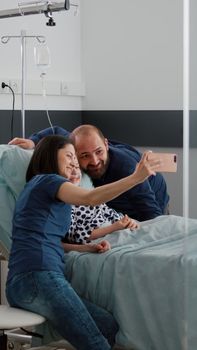 Happy family smiling while taking selfie using modern smartphone during medical examination in hospital ward. Little daughter wearing oxygen nasal tube recovering after disease surgery