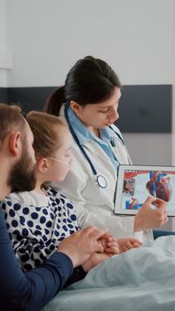 Pediatric cardiologist woman doctor explaining heart illness using tablet with medical cardiogram on display during surgery consultation in hospital ward. Sick child recovering after sickness surgery