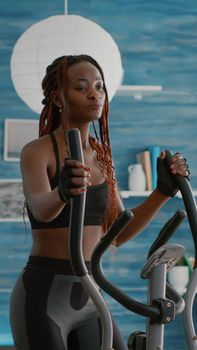 Fit athletic black woman running on elliptical bike watching wellness routine fitness video on tv during gym morning workout in living room. Athletic black woman training body muscle