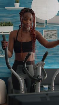 Black woman athletic training on elliptical bike practicing cardio sport watching aerobic video on tv during bodyweight morning routine in living room. Trainer enjoying healthy lifestyle