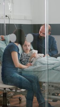 Worried parents sitting with sick girl daughter waiting for sickness expertise during medical examination in hospital ward. Child wearing oxygen nasal tube holding teedy bear while resting in bed