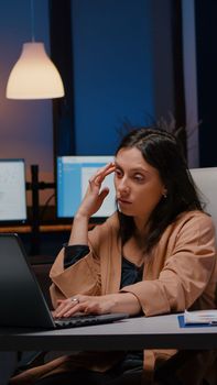 Overworked exhausted businesswoman working in startup office checking management strategy on laptop late at night. Stressed executive manager remains alone in company room after his collegue left