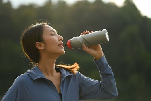 Woman drinking water from bottle, resting after jogging at the park in evening. Healthy lifestyle and fitness concept.