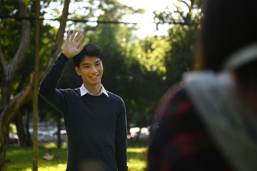 Friendly asian male student walking in campus and greeting his friend with hand raised up.