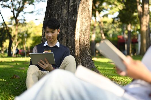 Smiling asian male students using tablet, preparing for exam while under tree in campus. Education and lifestyle concept.