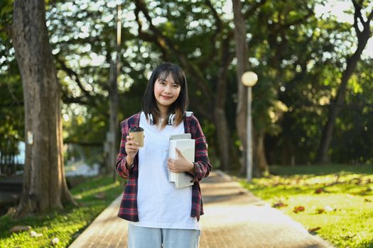 Portrait of happy asian female student holding books and paper cup in hands, going to lecture at the university.