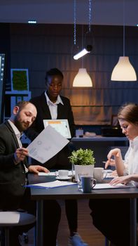 Focused workaholic entrepreneur woman with dark skin explaining management strategy using tablet. Business diverse multi-ethnic teamwork overworking in company office meeting room late at night