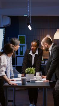 Diverse multi ethnic businesspeople discussing over graphs paperwork standing at conference table overworking in company meeting room. Teamwork brainstorming presentation ideas late at night