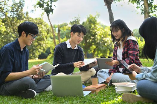 Group of friends, university students reading book, preparing for exam on green lawn at campus.