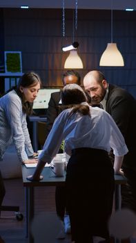 Multi ethnic businesspeople teamwork standing at conference table analyzing management graphs in evening. Diverse teamwork discussing company presentation in meeting room while brainstorming