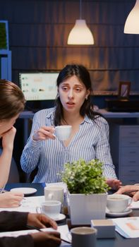 Businesswoman holding cup of coffee while discussing with multi-ethnic teamwork solving management project using graphs paperwork. Diverse coworkers working in meeting room late at night