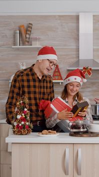 Grandparents giving wrapper xmas present gift with ribbon on it to grandchild standing at table in decorated kitchen. Happy family enjoying winter season celebrating christmas holiday