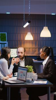 Manager woman writing management statistics analyzing graphs presentation on computer while diverse teamwork brainstorming company ideas. Multi-ethnic businesspeople working in meeting room in evening