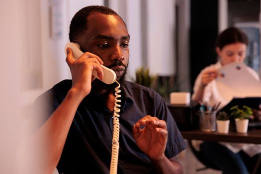 Project manager discussing startup strategy plan on landline phone, african american employee answering colleague call. Businessman having telephone communication in coworking space