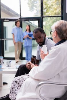 Specialist medic showing painkiller bottle to african american patient while explaining health care treatment during checkup visit appointment in hospital lobby. Medicine service and concept