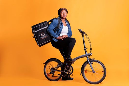 Pizzeria deliverywoman carrying food takeaway backpack while riding bicycle in studio with yellow background, delivering pizza order for dinner. Food transportation service and takeaway concept