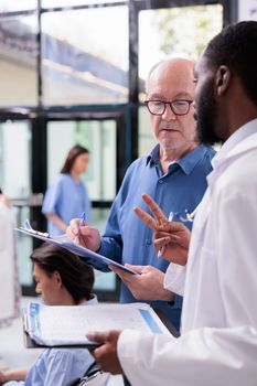 Specialist doctor discussing disease diagnosis to senior patient during checkup visit consultation in hospital reception. Medic explaining healthcare treatment while man signing medical report