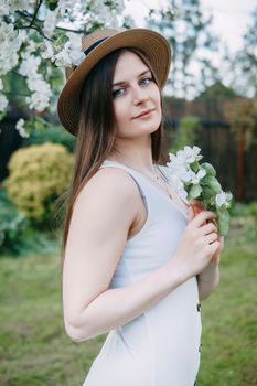 Beautiful young girl in white dress and hat in blooming Apple orchard. Blooming Apple trees with white flowers
