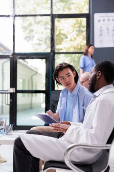 Physician helping asian patient to sign medical insurance after explaining disease expertise during checkup visit consultation in hospital waiting area. Diverse people standing in reception