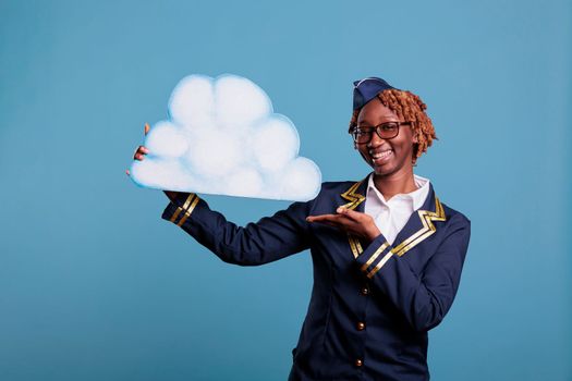 Female flight attendant smiling while holding paper cloud against blue background in studio shot. Optimistic woman stewardess wearing uniform, concept of flying among the clouds.