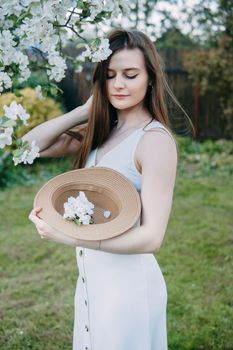 Beautiful young girl in white dress and hat in blooming Apple orchard. Blooming Apple trees with white flowers