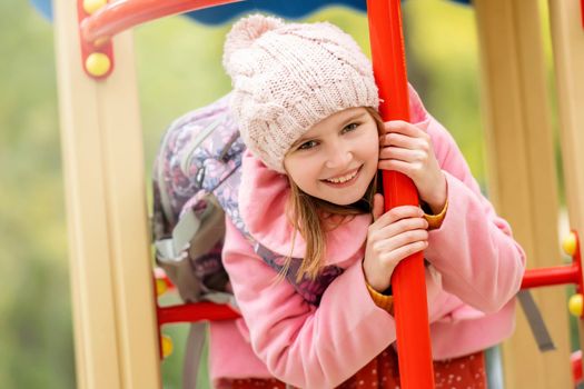 Pretty girl kid playing on playground at autumn day outdoors, looking at camera and smiling. Female child portrait at street