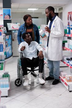 African american pharmacist helping man in wheelchair choosing prescription medications. Pharmacy consultant recommending medicaments to client, customer with disability and social worker in drugstore