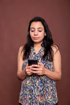 Serious indian woman chatting in social networks, using smartphone, sending sms. Person holding mobile phone, talking online, messaging on telephone, front view studio medium shot