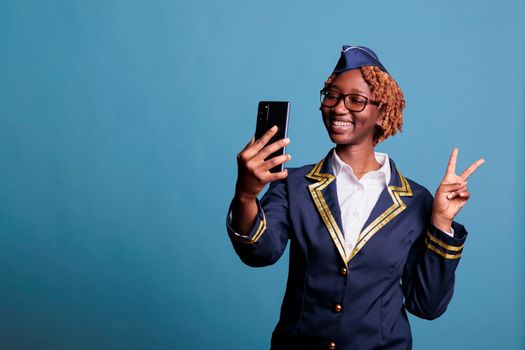 Funny female flight attendant on video call with friends during break time at work. Stewardess dressed in uniform using mobile phone while having fun chatting, studio shot.