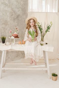 A girl with long hair in a dress and hat is sitting at the Easter table with cakes, spring flowers and quail eggs. Happy Easter celebration