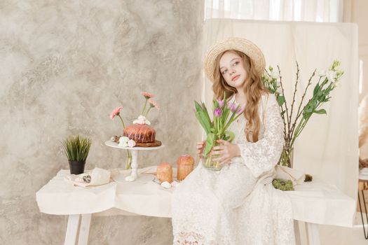 A girl with long hair in a dress and hat is sitting at the Easter table with cakes, spring flowers and quail eggs. Happy Easter celebration