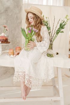 A girl with long hair in a dress and hat is sitting at the Easter table with cakes, spring flowers and quail eggs. Happy Easter celebration