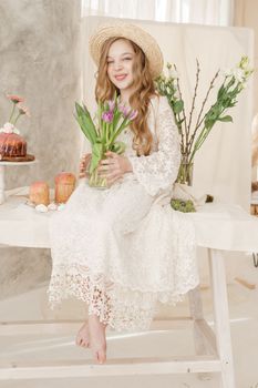 A girl with long hair in a dress and hat is sitting at the Easter table with cakes, spring flowers and quail eggs. Happy Easter celebration