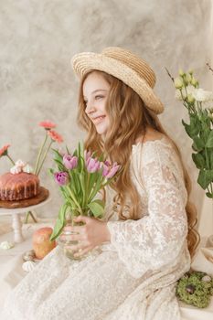 A girl with long hair in a dress and hat is sitting at the Easter table with cakes, spring flowers and quail eggs. Happy Easter celebration