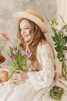 A girl with long hair in a dress and hat is sitting at the Easter table with cakes, spring flowers and quail eggs. Happy Easter celebration