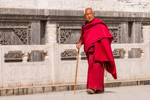 Tibetan monk in red robe with stick walks along a decorated wall of Kumbum Champa Ling Monastery, Xining, China