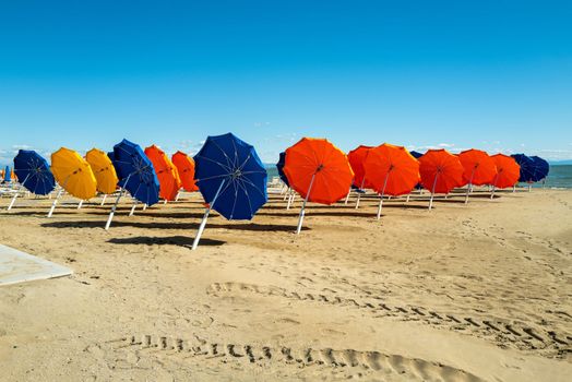 A large collection of colorful umbrellas await guests on a sandy beach in bright blue skies and sunshine.