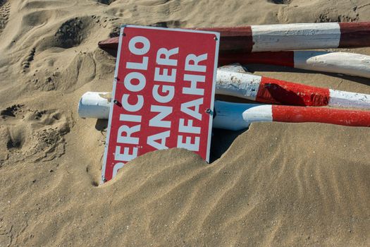 In the fine sand on the sea beach lies a sign with the inscription "Danger" in three different languages.