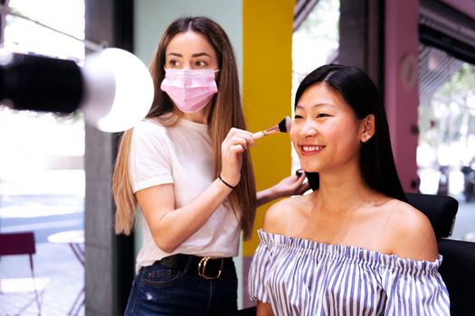 professional make-up artist doing makeup to a smiling young woman in front of the mirror of a beauty salon, concept of wellness and body care