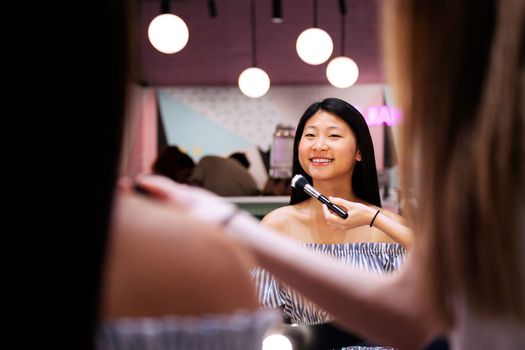 professional makeup artist applying make-up with a brush to a smiling young woman in front of the mirror in the beauty salon, concept of wellness and body care