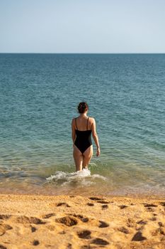 Woman sea swimsuit sand. A girl in full growth stands with her back and enters the sea in a black swimsuit. Alone on the beach on a sunny day