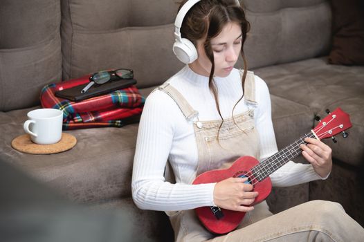 Beautiful young hipster girl in casual clothes learning to play the ukulele guitar while sitting on the floor in the living room at home, hobby leisure time concept.