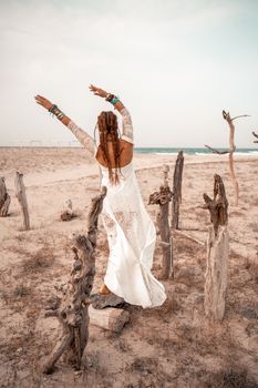 Model in boho style in a white long dress and silver jewelry on the beach. Her hair is braided, and there are many bracelets on her arms