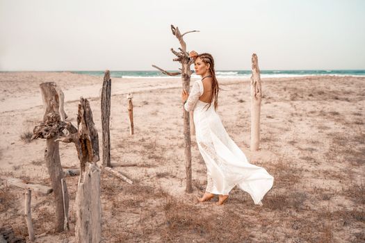 Model in boho style in a white long dress and silver jewelry on the beach. Her hair is braided, and there are many bracelets on her arms