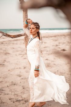 Model in boho style in a white long dress and silver jewelry on the beach. Her hair is braided, and there are many bracelets on her arms